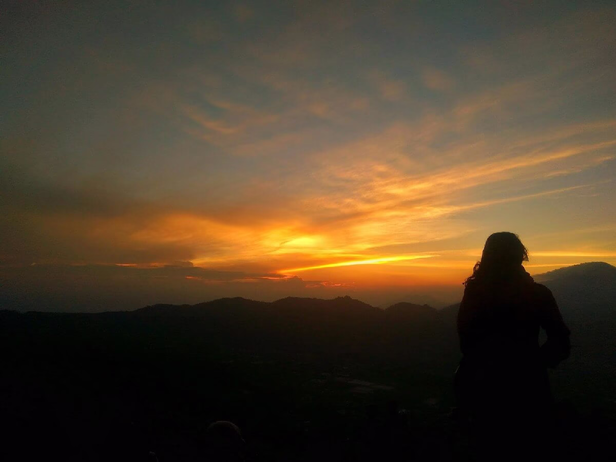 Silhouette seseorang menikmati matahari terbenam di pegunungan, menciptakan suasana damai dan reflektif.
