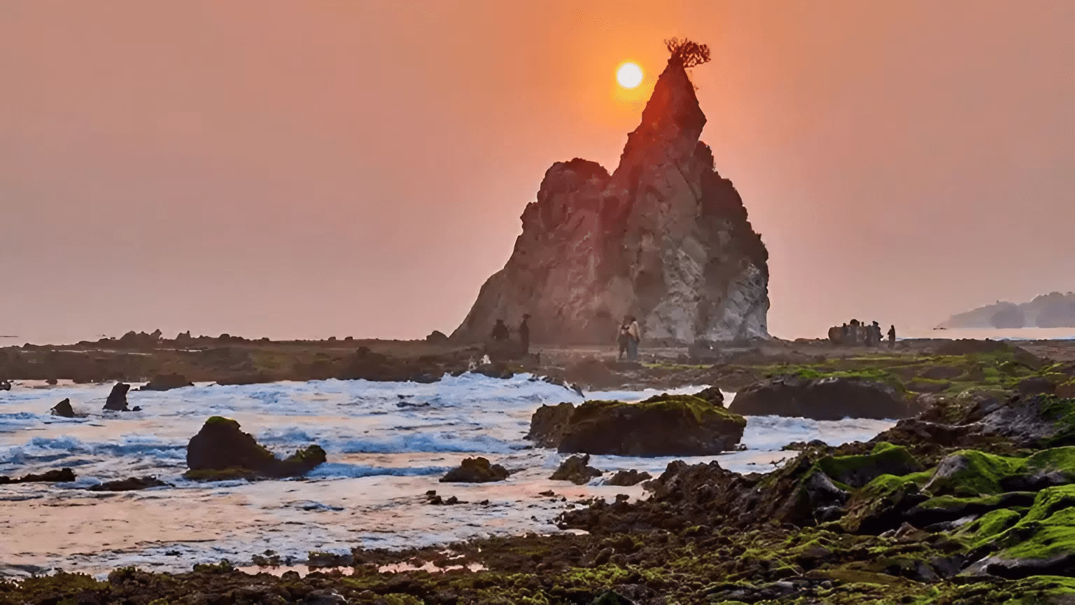 Pemandangan matahari terbenam di pantai dengan batu besar dan pengunjung menikmati keindahan alam yang menenangkan.