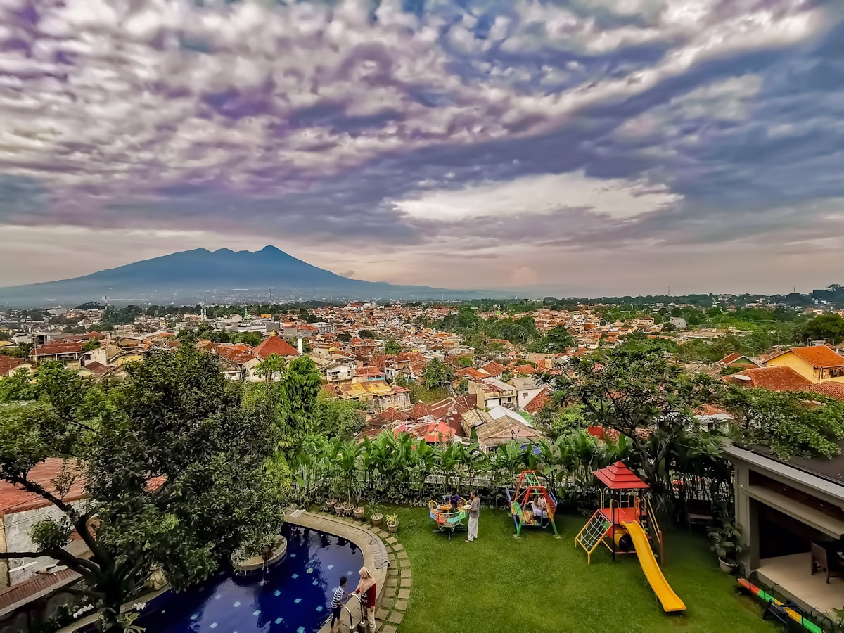 Pemandangan kota dengan gunung di latar belakang, taman bermain ramah anak, dan kolam renang di halaman rumah.