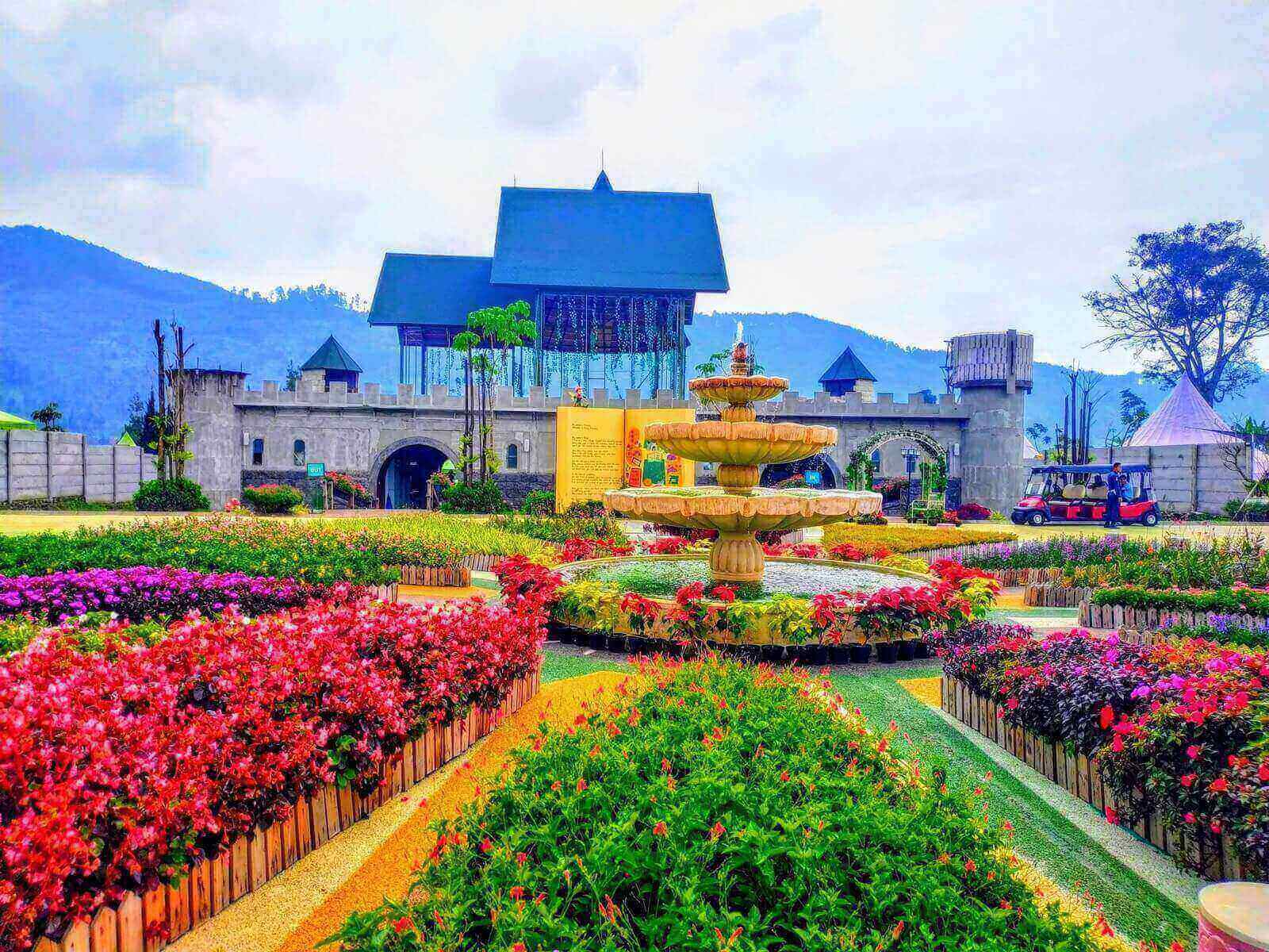 Taman indah dengan air mancur dan bunga colorful menghiasi latar belakang kastil, menciptakan suasana damai dan menarik.