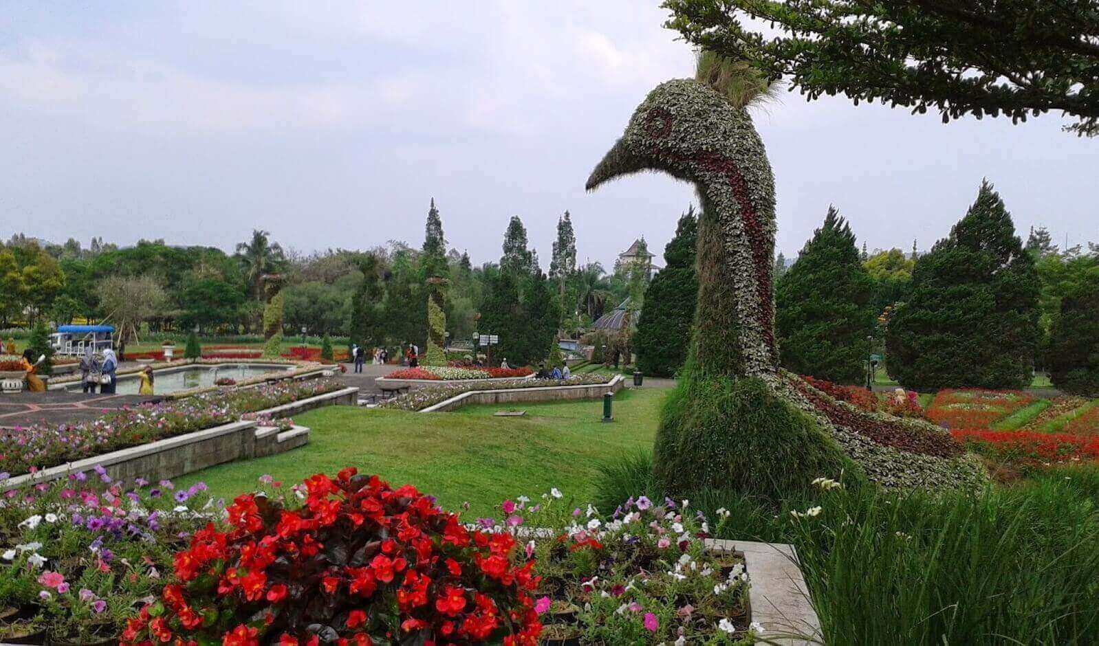 Taman indah dengan patung burung hias dari tanaman, dikelilingi bunga berwarna-warni dan pengunjung menikmati suasana.