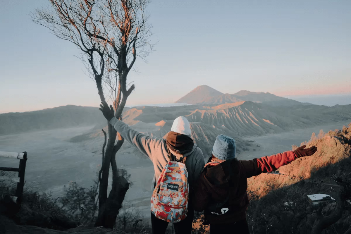 Dua wisatawan berdiri di puncak bukit menikmati pemandangan indah Gunung Bromo saat matahari terbit, merasakan kebebasan.