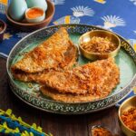 "Piring hidangan rendang tradisional dengan keripik dan telur setengah matang, dikelilingi oleh rempah-rempah berwarna cerah."