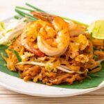 Pad Thai Thailand dengan udang, tauge, dan irisan limau, disajikan di atas daun pisang segar.