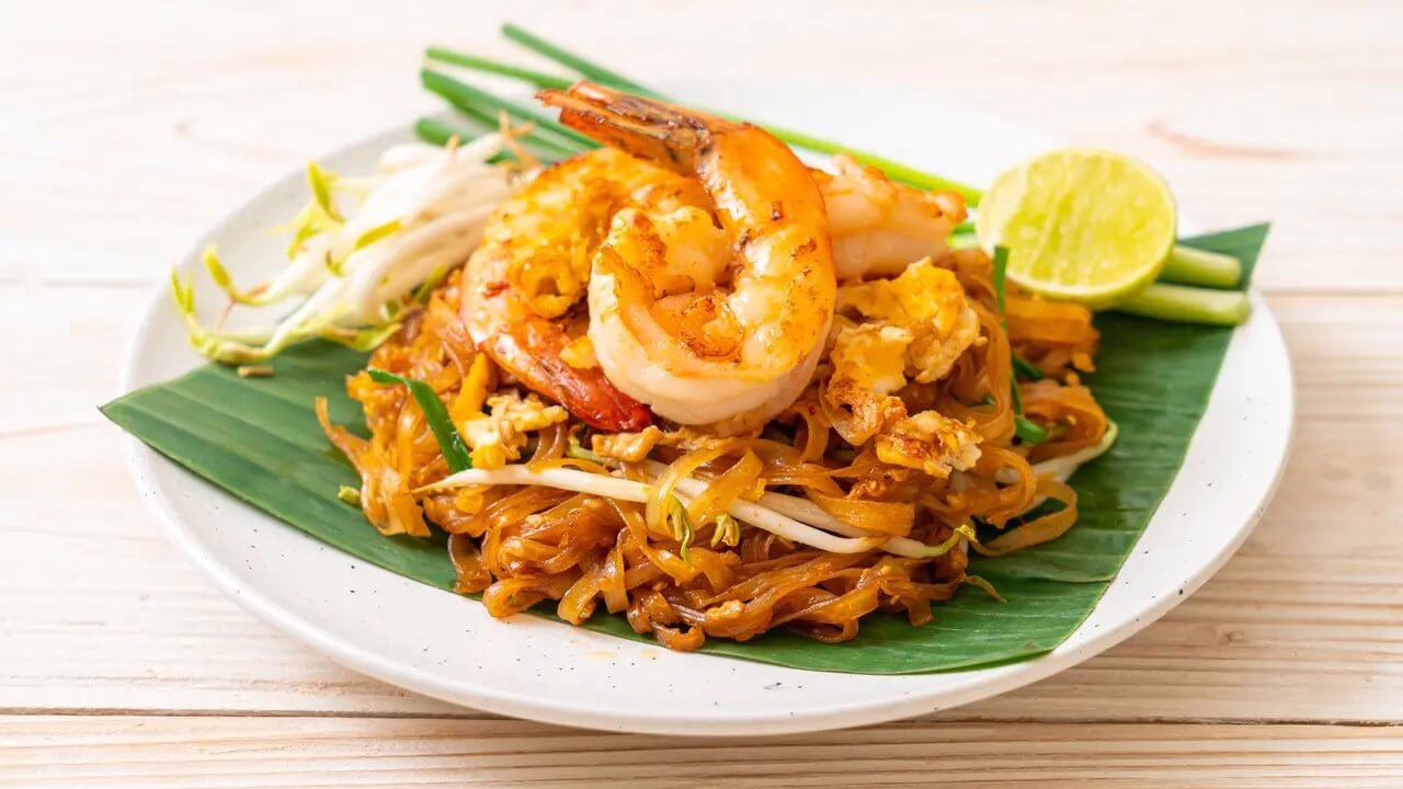 Pad Thai Thailand dengan udang, tauge, dan irisan limau, disajikan di atas daun pisang segar.