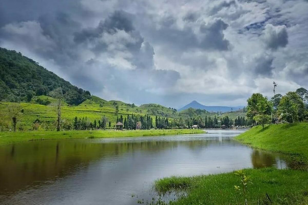 Pemandangan tenang dan hijau di tepi danau, dikelilingi perbukitan dan langit berawan yang menambah keindahan alam.