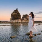 Kannaya Nareswari mengenakan gaun putih berdiri di tepi pantai berbatu saat matahari terbenam, menciptakan suasana tenang.