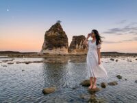 Kannaya Nareswari mengenakan gaun putih berdiri di tepi pantai berbatu saat matahari terbenam, menciptakan suasana tenang.